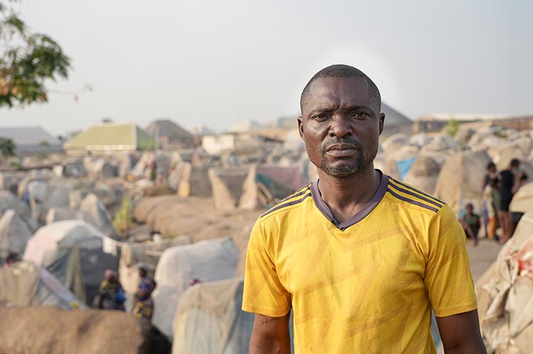 Pastor Barnabas steht vor einer Zeltstadt. Diese dient als Flüchtlingsunterkunft für verfolgte Geschwister aus Nigeria.