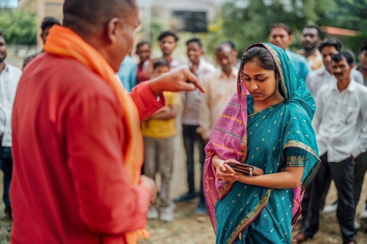 Eine indische junge Christen hält eine Bibel in ihren Händen. Ein Geistlicher in einem orangefarbenem Gewand betet für sie.Im Hintergrund sind verschwommen weitere Personen zu sehen. 