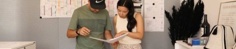 Ein junger Mann mit Kappe und eine asiatische Frau stehen vor einem Kalender und planen etwas auf einem Papier