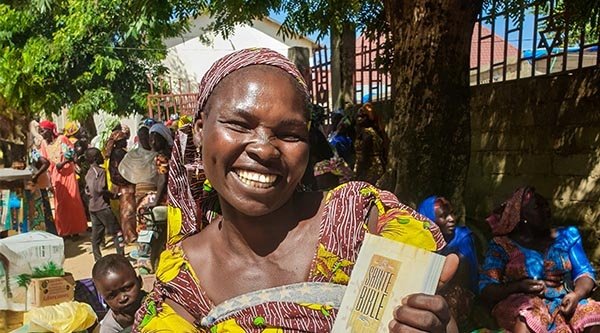 Eine afrikanische Christin präsentiert lächelnd ihre Bibel. Im Hintergrund sind zahlreiche Menschen aus ihrer Gemeinschaft.