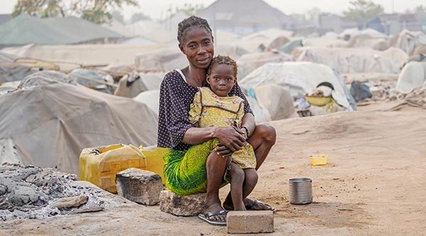 Eine Frau sitzt mit ihrer kleinen Tochter auf einem Stein. Im Hintergrund sind die Zelte eines Flüchtlingslagers in Nigeria zu sehen.